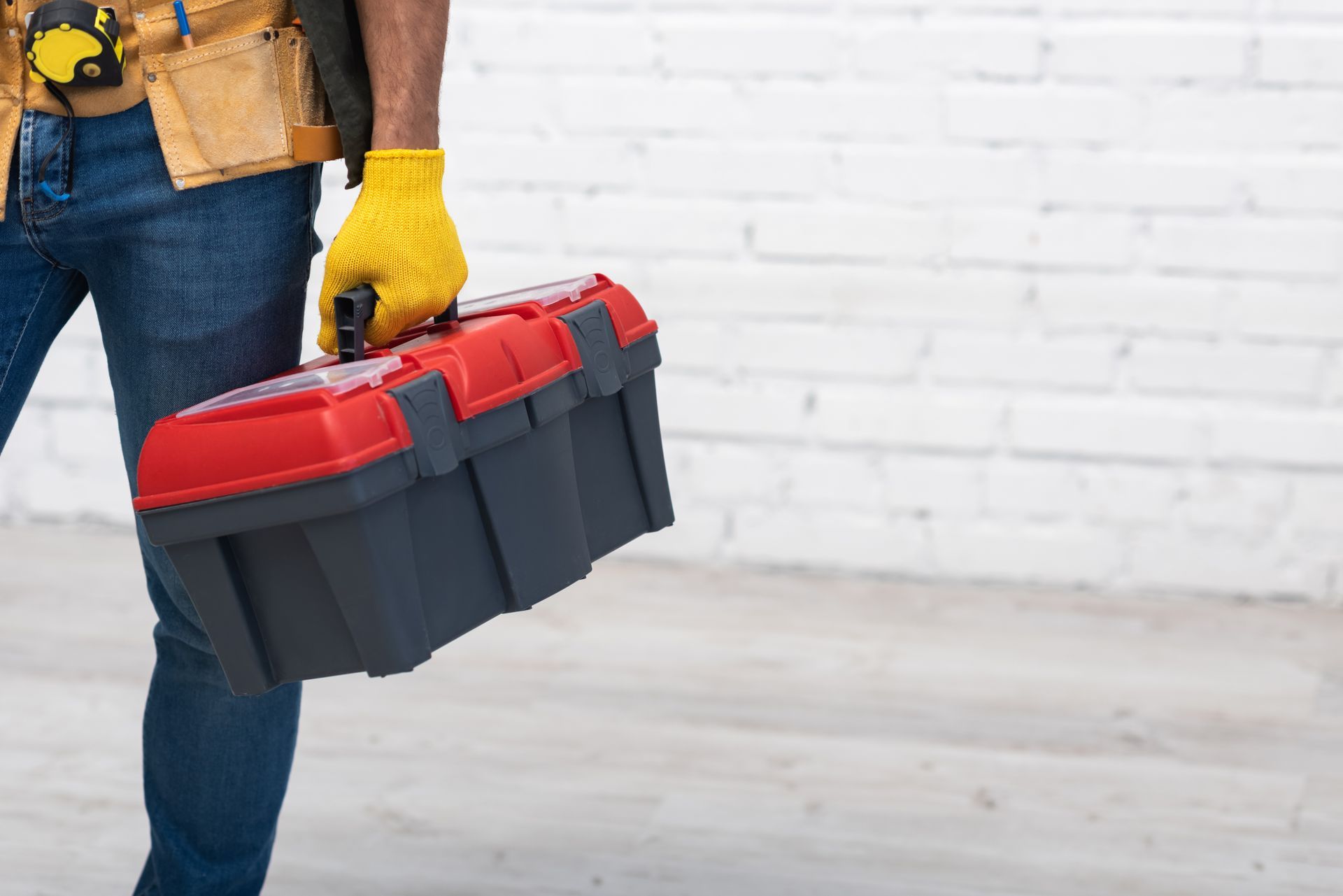 Un ouvrier tenant une boîte à outils rouge et noire, portant des gants jaunes et un jean, se tient devant un mur blanc.