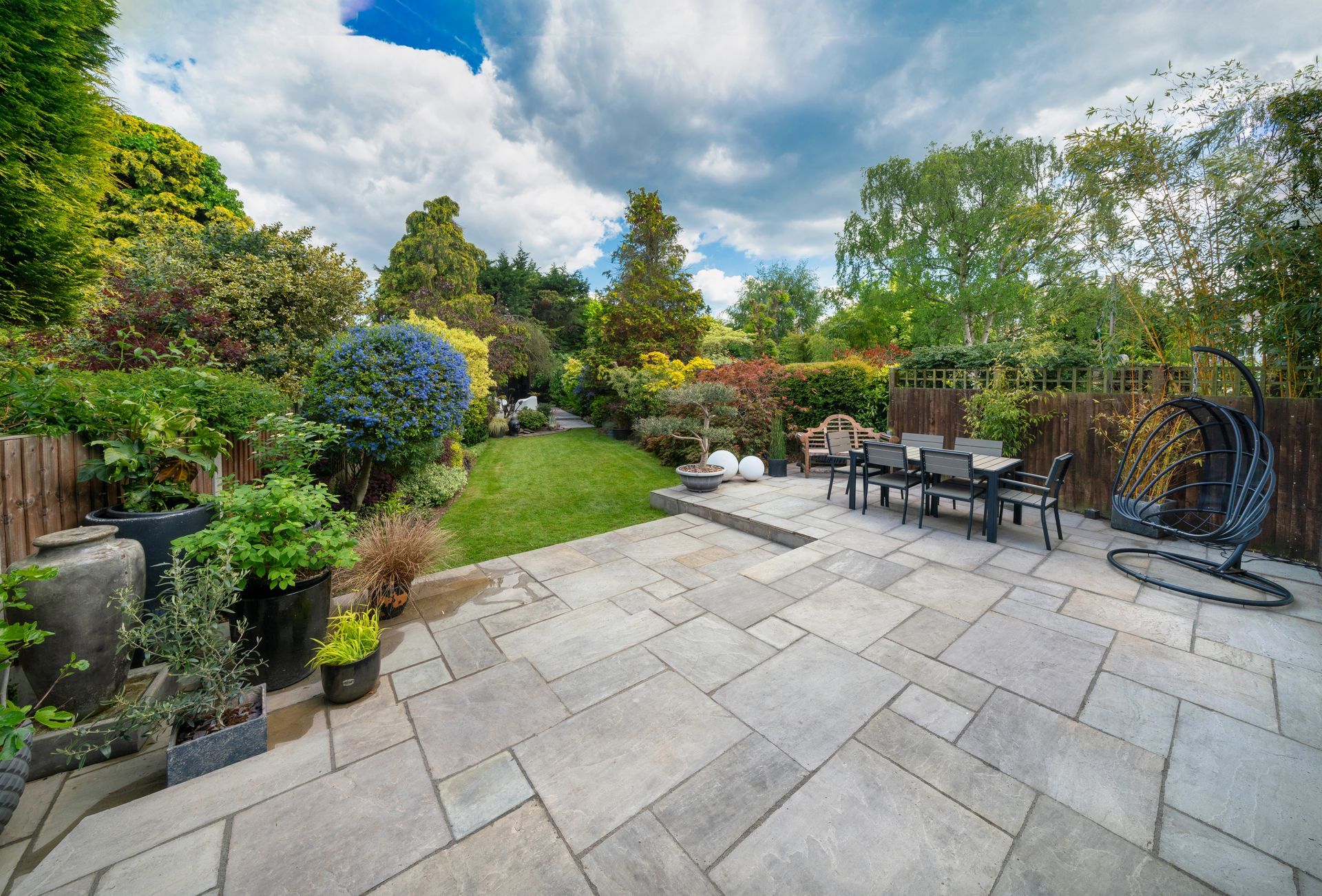 Terrasse arrière avec pavage en pierre, sièges extérieurs, végétation luxuriante et petite pelouse sous un ciel nuageux.