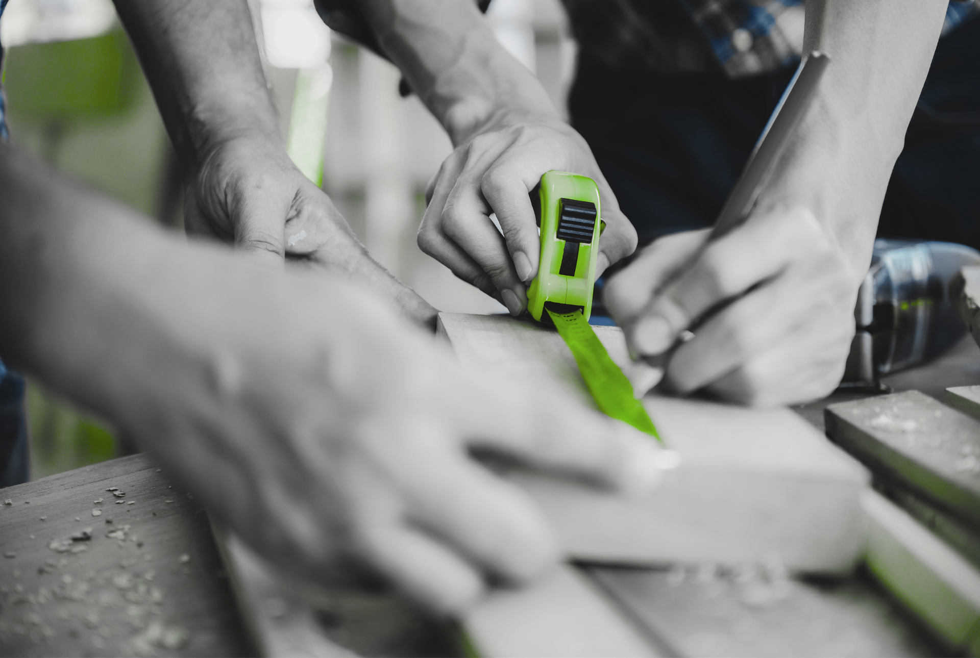 Des mains utilisent un mètre ruban vert sur un établi avec des morceaux de bois dans un atelier.