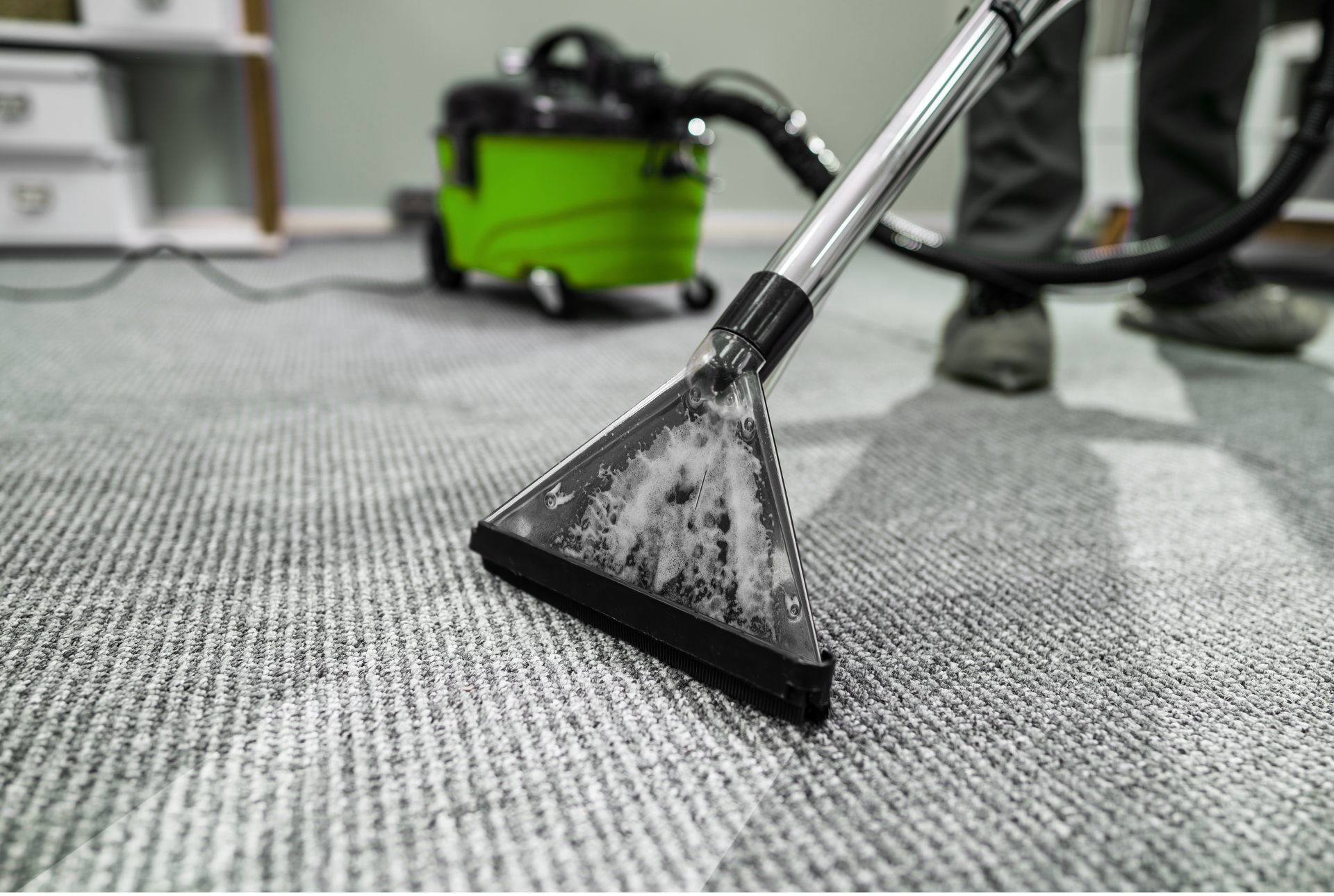 Buse d'aspirateur pour tapis passant l'aspirateur sur un tapis gris dans une pièce, avec une machine de nettoyage verte en arrière-plan.