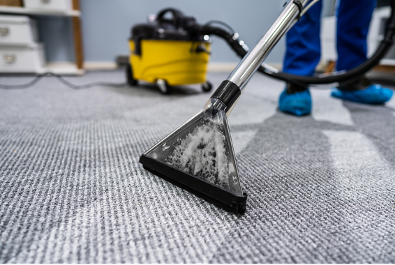 Buse d'un nettoyeur de tapis nettoyant un tapis gris dans une pièce où l'on aperçoit un aspirateur jaune en arrière-plan.