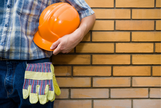 Ouvrier du bâtiment tenant un casque orange et des gants jaunes contre un mur de briques.