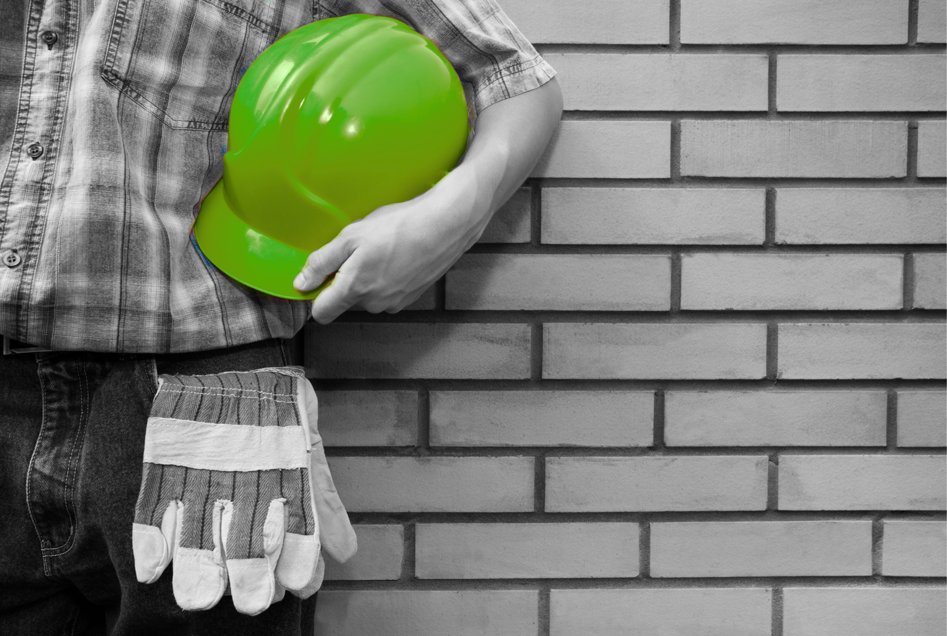 Ouvrier tenant un casque vert et des gants contre un mur de briques.