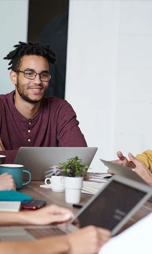 Un hombre con gafas sonríe durante una reunión, con una computadora portátil, una taza y una planta sobre la mesa.