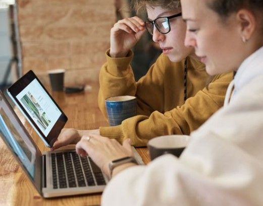 Dos personas utilizan computadoras portátiles, una de ellas mira una tableta, en una mesa de madera en un café.