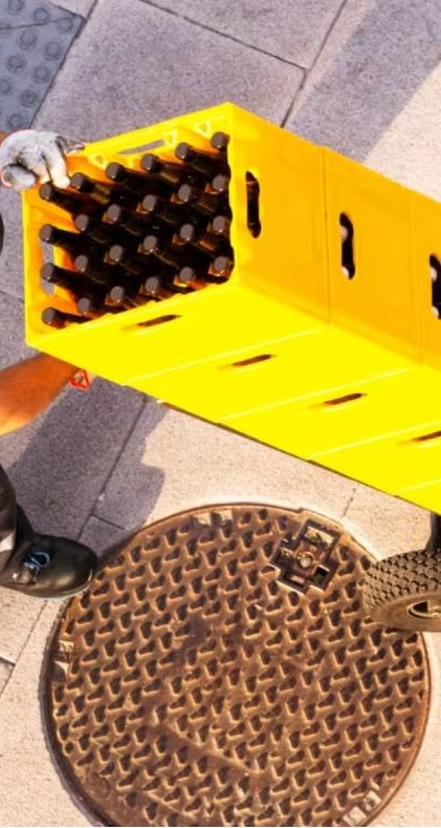 Eine Person in schwarzer Kleidung und Kappe trägt einen gelben Kasten mit dunklen Flaschen neben einem metallenen Kanaldeckel.