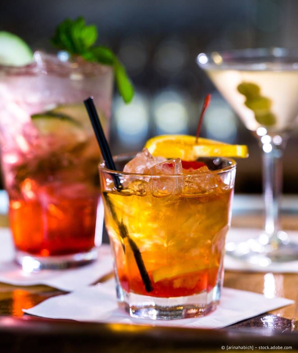 Cocktail auf einer Bar zusammen mit zwei anderen Getränken, garniert mit einer Orangenscheibe, einer Kirsche und einem schwarzen Strohhalm.