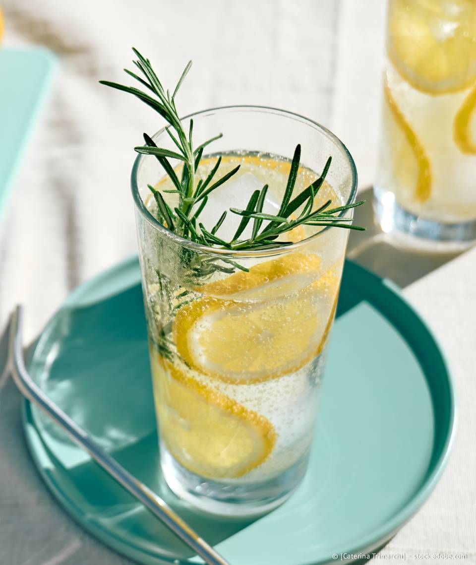 Ein Glas Limonade mit Zitronenscheiben, Eis und einem Rosmarinzweig auf einem türkisfarbenen Teller.
