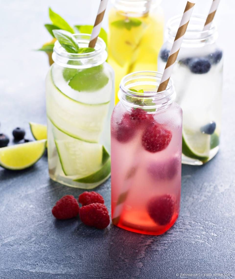 Vier Glasgefäße gefüllt mit buntem, aromatisiertem Wasser, mit Früchten und Kräutern, dazu Papierstrohhalme.
