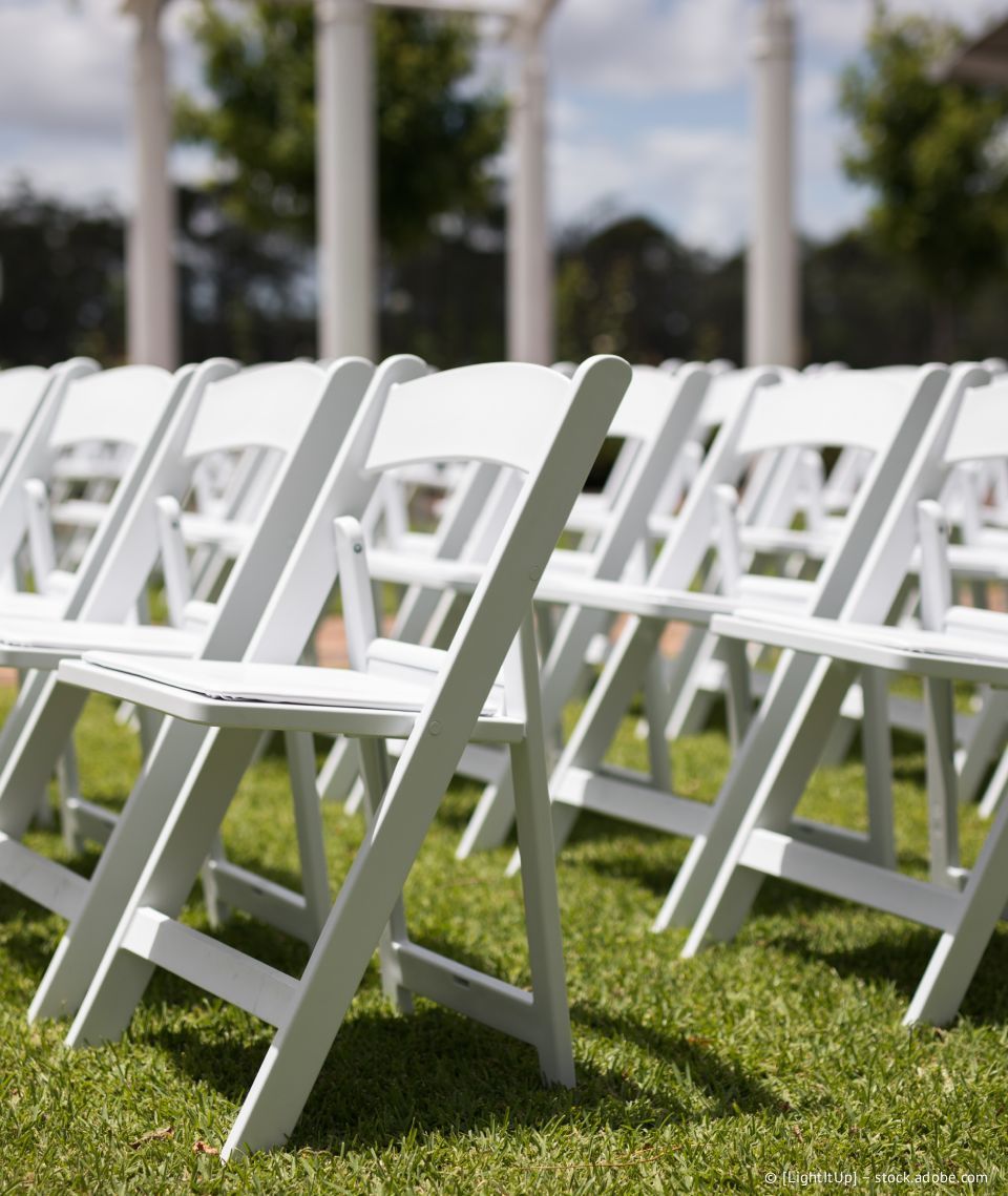 Weiße Klappstühle wurden für eine Veranstaltung im Freien auf einer Rasenfläche aufgestellt; im Hintergrund sind Säulen und Grünpflanzen zu sehen.