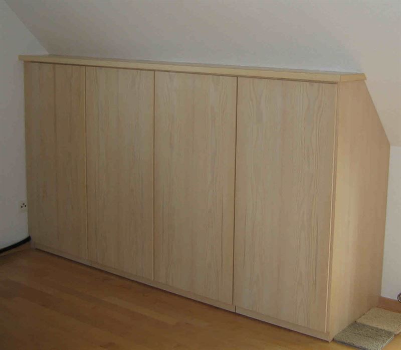 In einem Raum steht unter einer Dachschräge ein Holzschrank.