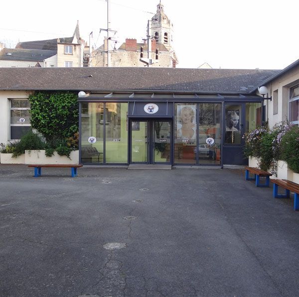 Entrée du musée avec portes vitrées, bancs bleus et bâtiment avec une tour en arrière-plan.