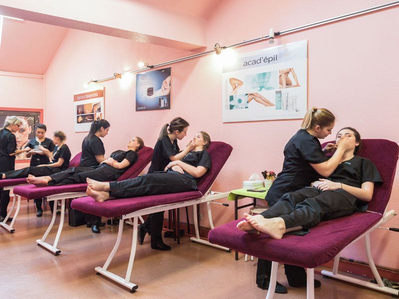 Ambiance salon de beauté, clients allongés sur des tables de massage pendant que d'autres leur prodiguent des soins. Murs roses, éclairage vif.