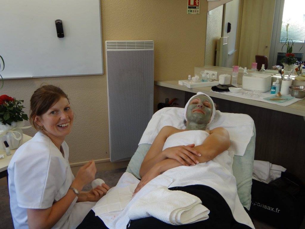 Une femme, le visage recouvert d'un masque de boue, se détend dans un spa ; l'esthéticienne sourit à ses côtés.