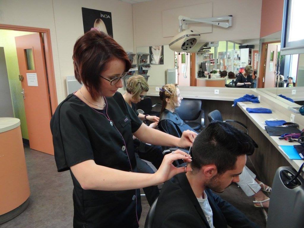 Dans un salon de coiffure, une coiffeuse aux cheveux foncés coupe les cheveux d'une cliente ; d'autres clients et coiffeurs sont visibles.