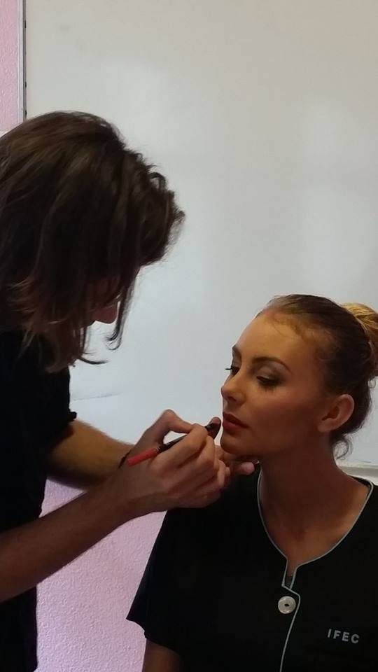Une maquilleuse applique du rouge à lèvres à une femme, devant un mur blanc.