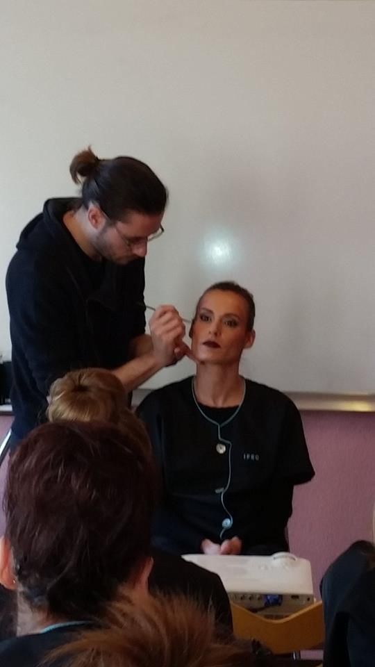Une maquilleuse applique du maquillage sur le visage d'une personne, probablement lors d'une démonstration. Des personnes observent la scène dans une salle de classe.