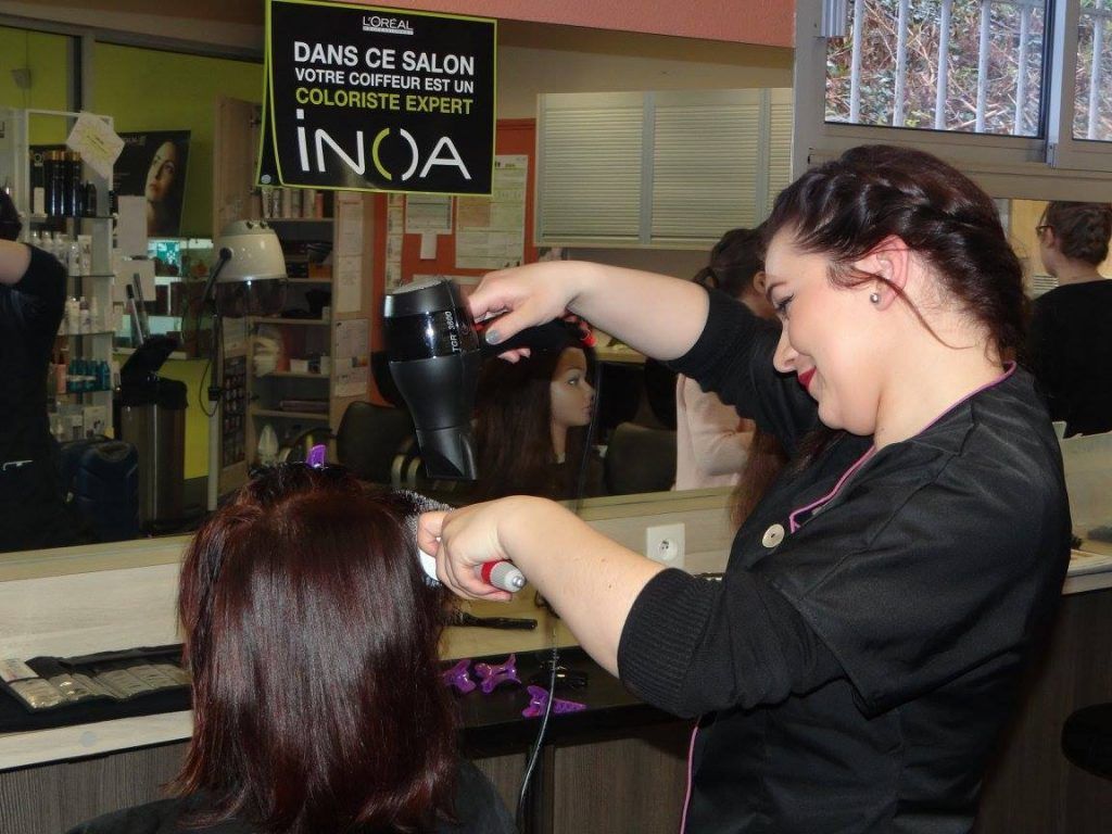 Dans un salon de coiffure, une coiffeuse sèche les cheveux d'une cliente aux cheveux roux foncé. La cliente sourit.