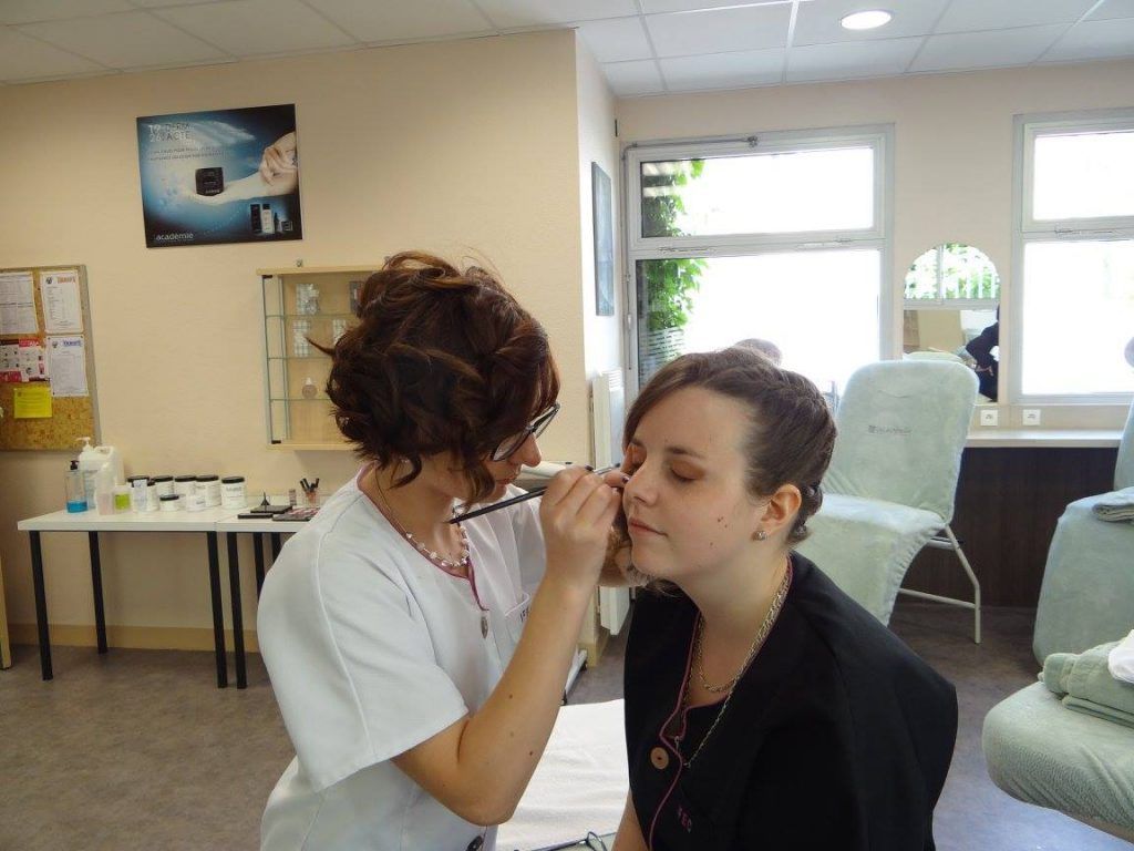 Une personne maquille les yeux d'une autre à l'intérieur. Une table et des chaises de maquillage sont visibles en arrière-plan.