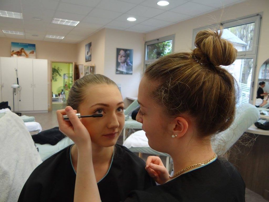 Une femme applique du mascara sur l'œil d'une autre. La scène se déroule dans un décor qui ressemble à un salon de beauté.