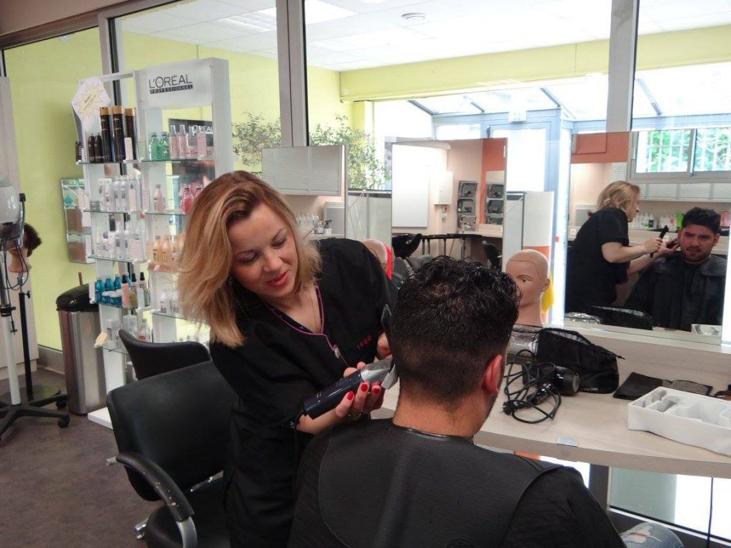 Une femme coupe les cheveux d'un homme dans un salon de coiffure ; une autre coiffeuse et un client sont visibles en arrière-plan.