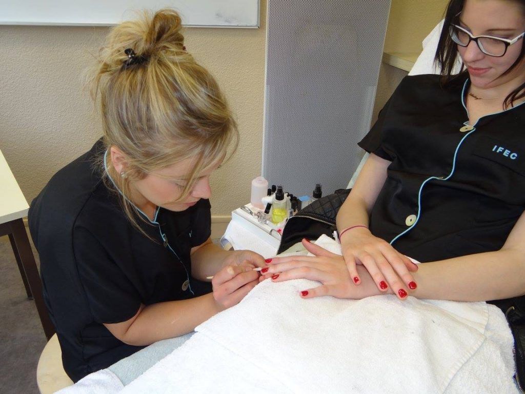 Une femme se fait faire une manucure dans un salon. La technicienne applique du vernis rouge sur les ongles de la cliente, assise sur une serviette blanche.