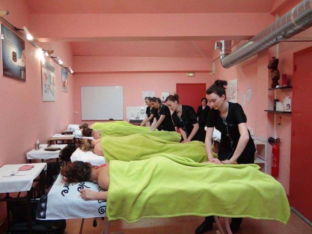 Cours de massothérapie : les étudiants s’exercent sur des clients. Des serviettes vertes recouvrent les tables de massage dans une salle rose.