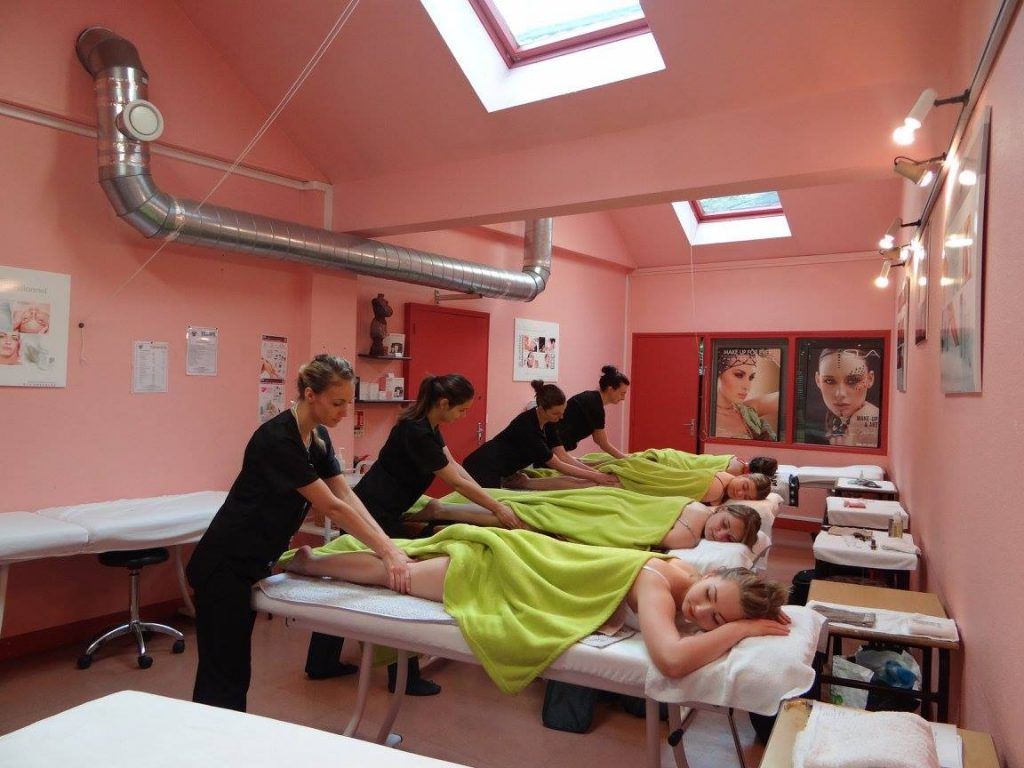 Des personnes reçoivent des massages des jambes dans une pièce rose. Les thérapeutes sont en tenue noire. Puits de lumière, serviettes vertes.