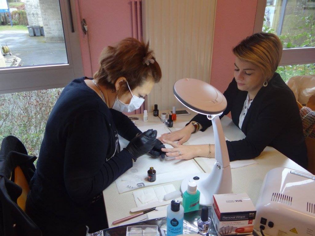 Une femme se fait faire une manucure à une table où sont disposés des produits de soin des ongles. Une technicienne applique du vernis sous une lumière artificielle.