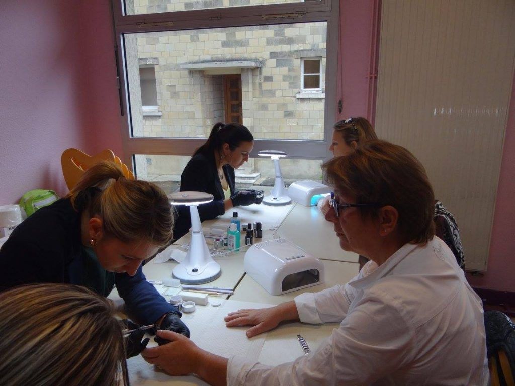 Des personnes travaillent à une table dans un salon de manucure. Il y a une fenêtre et les murs de la pièce sont roses.