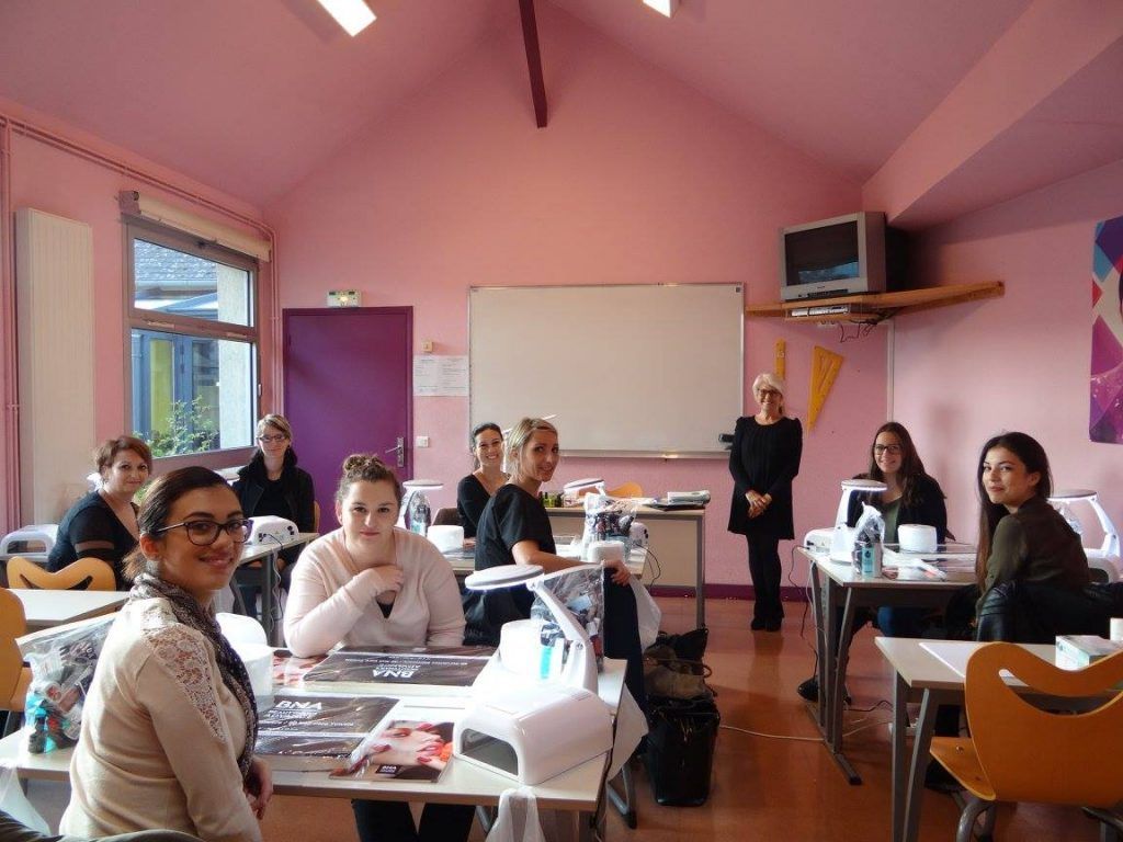 Une salle de classe avec des élèves et un professeur. Murs roses, tableau blanc et tables. Les élèves sont assis, certains sourient.