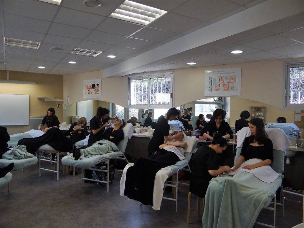 Dans une salle de classe d'une école d'esthétique, des personnes s'exercent à des soins sur des clientes allongées sur des tables de massage. Ambiance intérieure lumineuse.