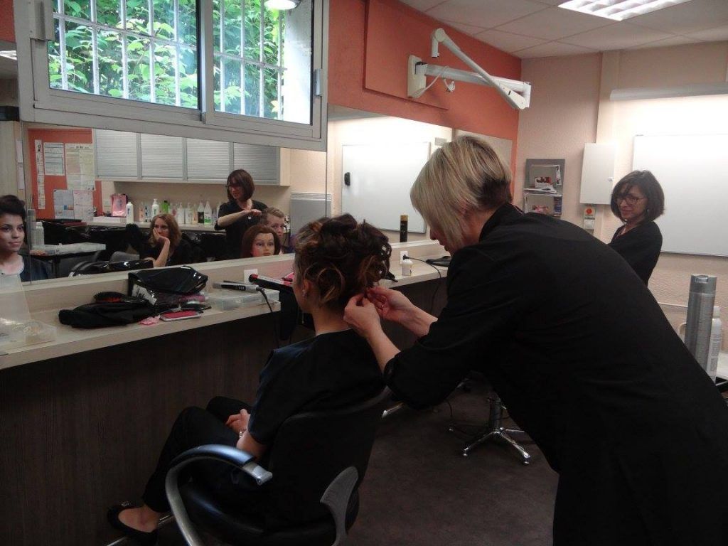 Une coiffeuse réalise une coiffure relevée sur une cliente dans un salon, en présence de plusieurs autres personnes.