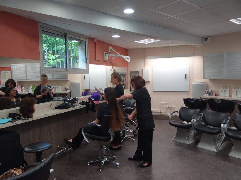 Cours de coiffure : Les élèves travaillent sur des mannequins et des clients dans un décor de salon de coiffure avec des postes de coiffage et des lavabos.