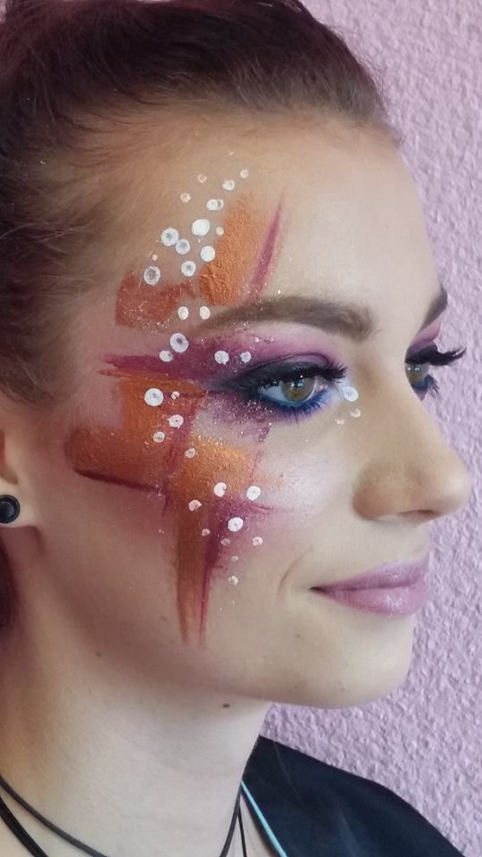 Femme au maquillage abstrait ; motif orange, violet et blanc sur le visage, longs cils, regardant à droite, souriante.