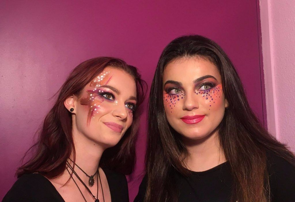 Deux femmes au maquillage original posent devant un mur rose. L'une a les cheveux roux, l'autre les cheveux bruns.