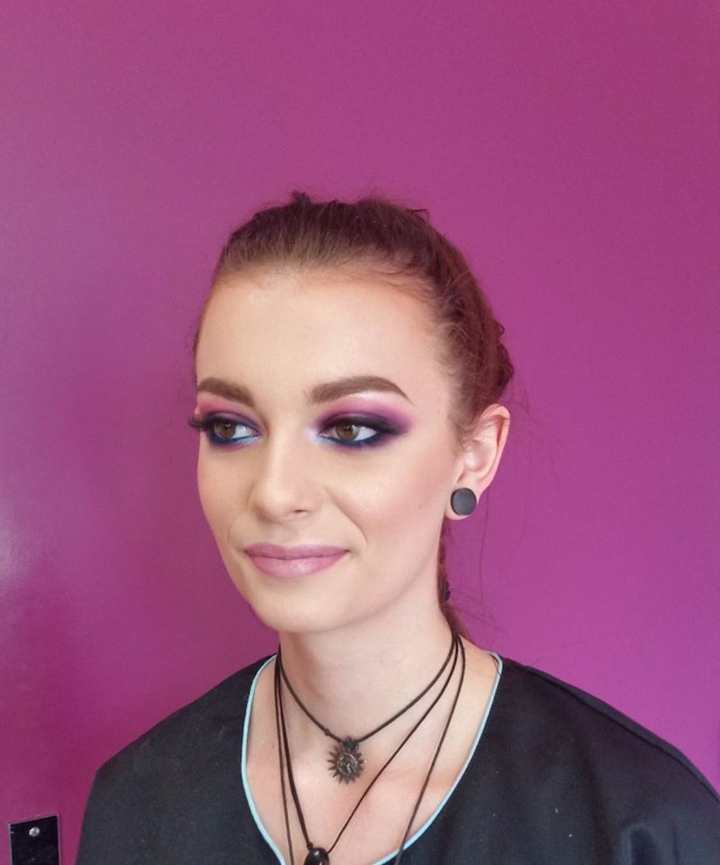 Femme au maquillage professionnel, fard à paupières foncé, fond rose. Porte plusieurs colliers.