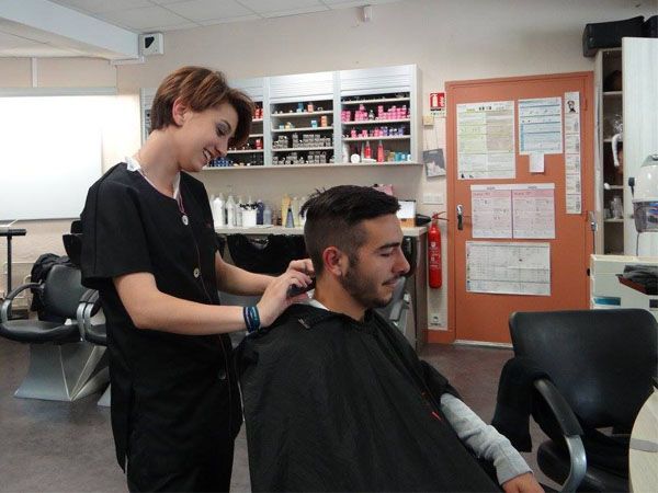 Un coiffeur en blouse noire coupe les cheveux d'un homme. Ambiance salon de coiffure, outils sur une étagère, homme portant une cape.