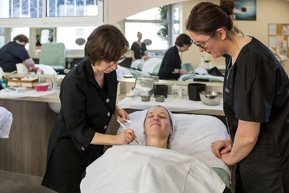 Deux personnes pratiquent des soins de la peau sur une cliente. Ambiance salon avec d'autres élèves qui travaillent.