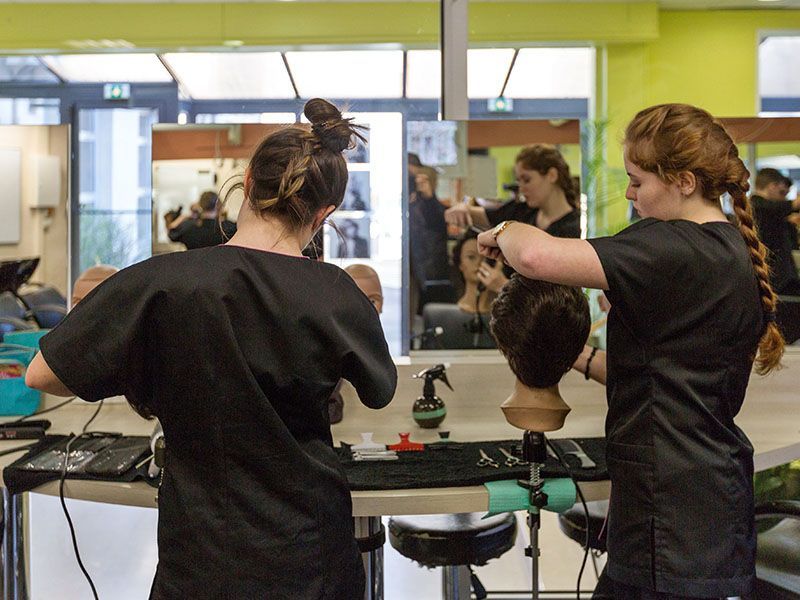Deux personnes vêtues de chemises noires s'entraînent à coiffer des têtes de mannequins à un poste de travail de salon.