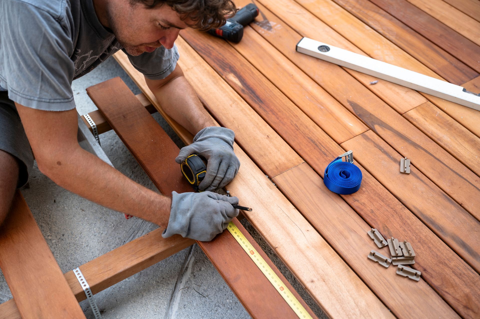 Un ouvrier mesure une planche de terrasse en bois, avec un mètre ruban et une perceuse électrique à proximité.