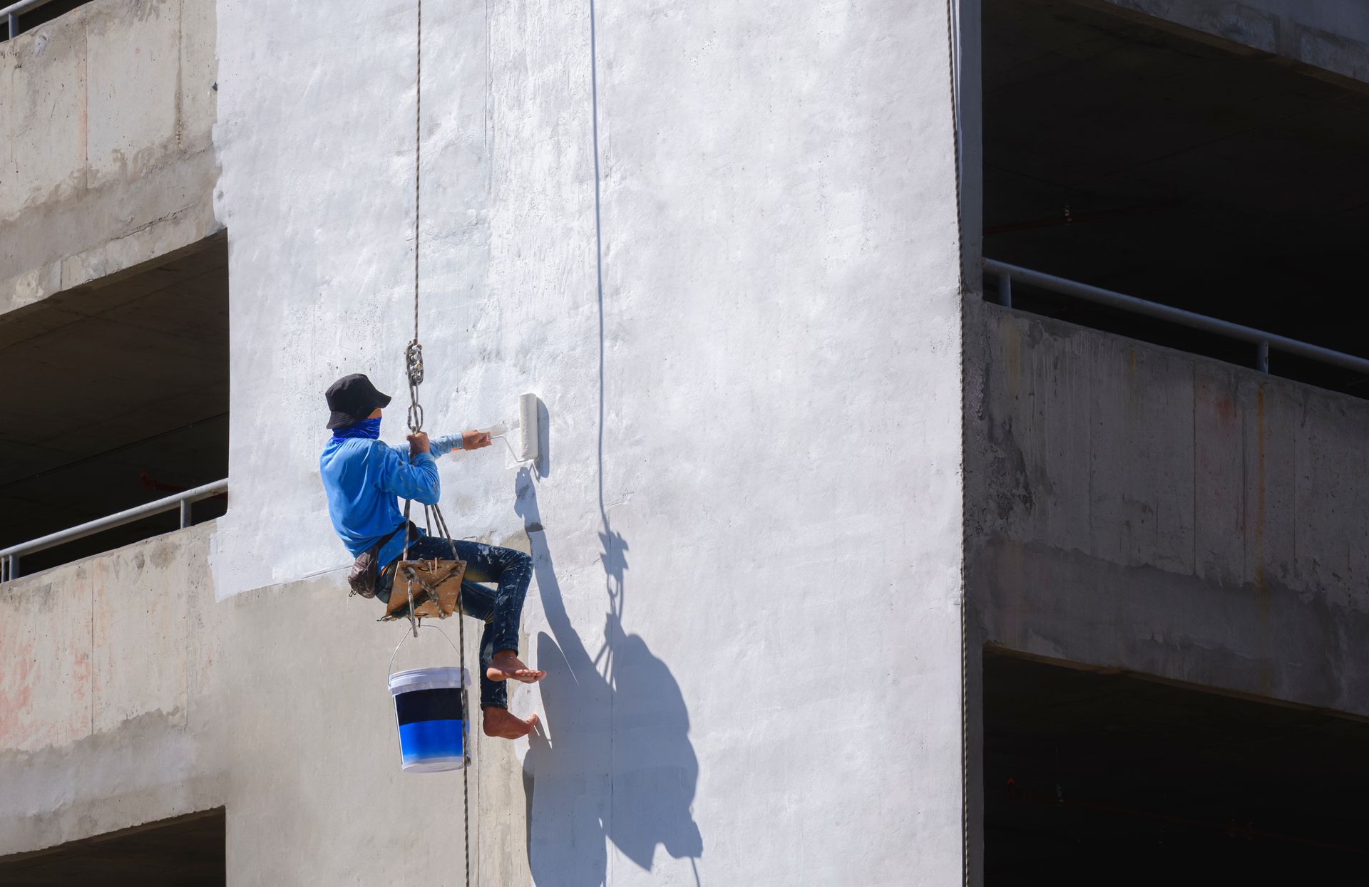Un ouvrier peint un haut mur blanc depuis une plateforme suspendue à l’extérieur d’un bâtiment