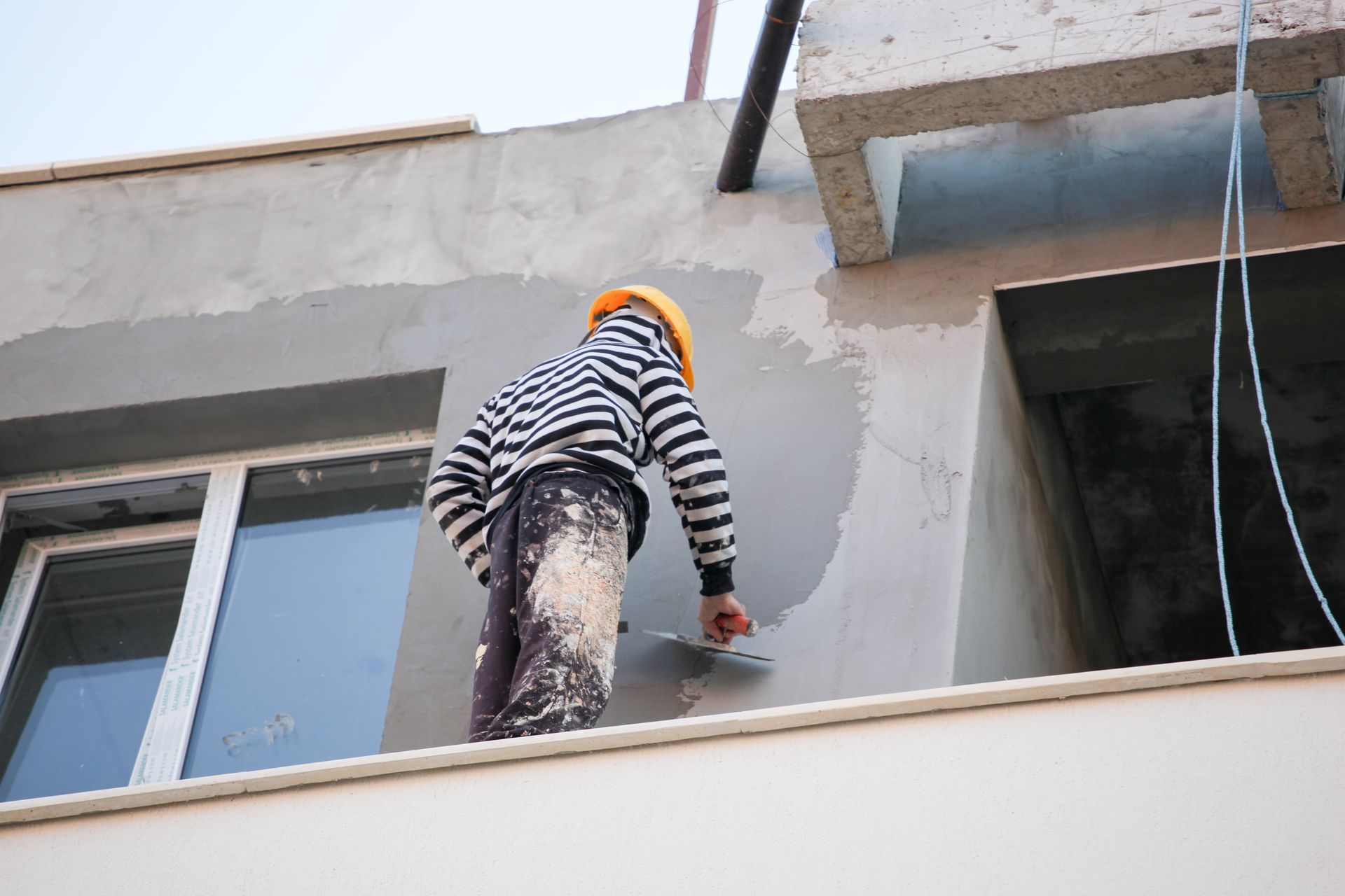 Un ouvrier en chemise rayée peint la façade extérieure d’un bâtiment depuis un balcon.