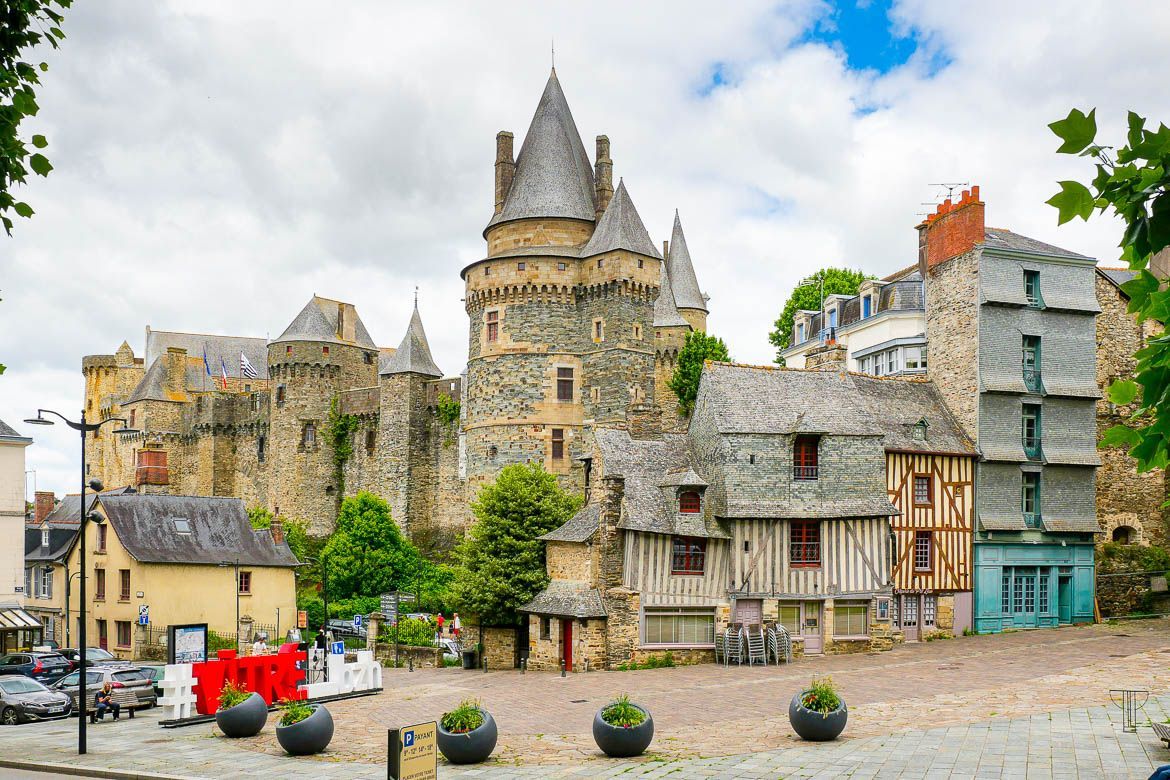 Château historique en pierre et maisons à colombages surplombant une place pavée, sous un ciel nuageux