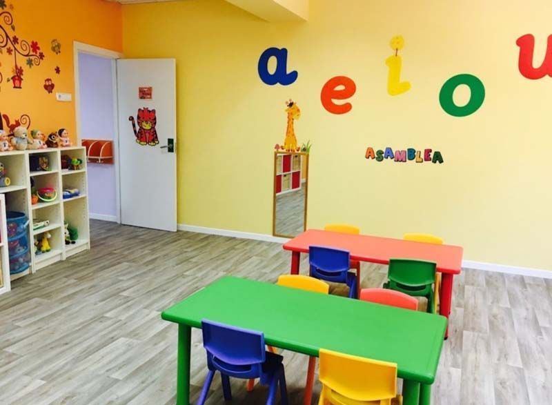Aula preescolar colorida con mesas y sillas, letras del alfabeto en la pared y juguetes en los estantes.