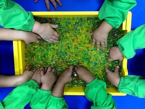 Manos de niños en un recipiente amarillo lleno de perlas de gel verdes y transparentes, con mangas verdes, sobre una superficie azul.