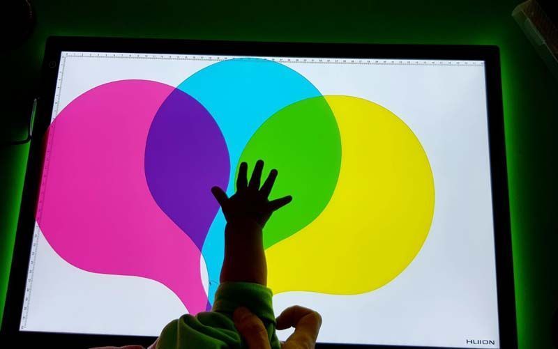 La mano de un niño se extiende hacia un panel iluminado con círculos superpuestos de color rosa, azul, verde y amarillo, mostrando la mezcla de colores.