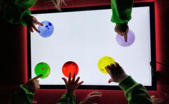 Manos de niños en un recipiente amarillo lleno de perlas de gel verdes y transparentes, con mangas verdes, sobre una superficie azul.
