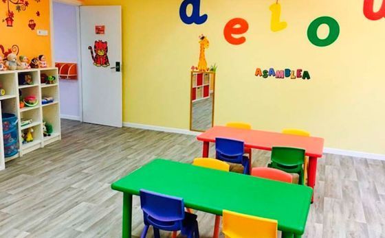 Aula preescolar colorida con mesas, sillas, estantes y decoraciones de letras en la pared.