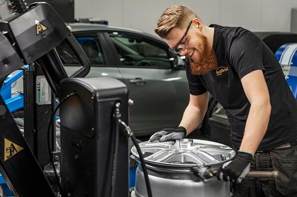 Un mecánico trabaja en una rueda de coche, sonriendo, en un taller. Se ve un coche gris y equipo azul.